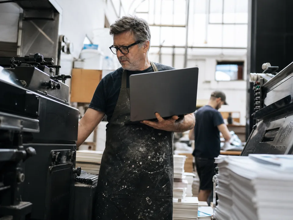 Printing business owner holding a laptop between printing machines